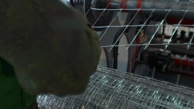 A close-up view of a person inspecting a large roll of galvanized wire mesh in an industrial setting. The person's head and shoulder are visible in the foreground on the left, while their gloved hands