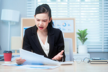 An Asian woman in a formal black suit sits at her office desk, holding documents with a tense...