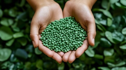 Hands holding a pile of small, green, fertilizer pellets against a backdrop of lush green leaves.