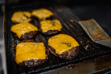 Juicy beef burger patties topped with melted cheddar cheese sizzling on a hot grill, with grease and a metal spatula nearby.