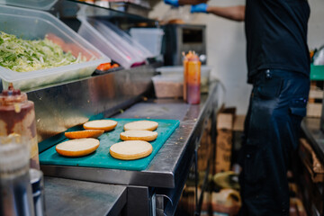 Burger buns are laid out on a green cutting board in a commercial kitchen, with a worker preparing...