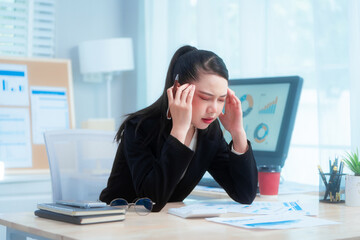 An Asian woman in a formal black suit sits at her office desk, holding documents with a tense...