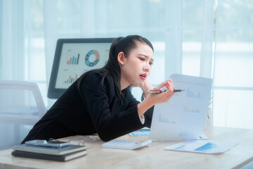 An Asian woman in a formal black suit sits at her office desk, holding documents with a tense...