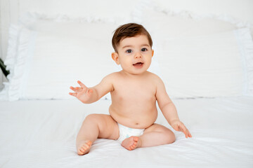 baby in diapers on a white bed at home, space for text, little child boy smiling on the bed before...