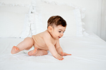 baby in diapers on a white bed at home, space for text, little child boy smiling on the bed before...