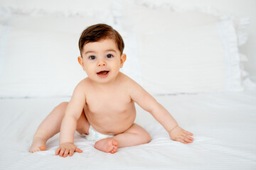 baby in diapers on a white bed at home, space for text, little child boy smiling on the bed before...