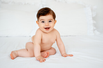 baby in diapers on a white bed at home, space for text, little child boy smiling on the bed before...