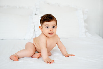 baby in diapers on a white bed at home, space for text, little child boy smiling on the bed before...