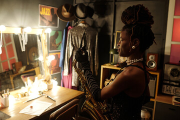 Black woman young adult holding saxophone standing in dressing room preparing for jazz performance, musical instruments and stage costumes visible in background, looking away from camera
