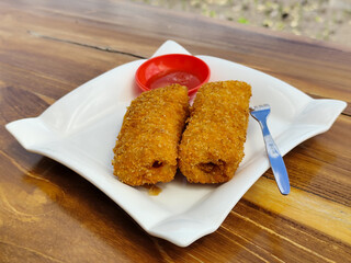 Crispy golden rissoles with savory and delicious filling, served on an elegant white plate, accompanied by a small bowl filled with bright red chili sauce and a fork, on the menu at an outdoor cafe.