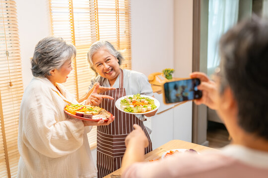 Group of Happy Asian senior woman having dinner together at home. Elderly retired women friends using mobile phone taking picture during preparing vegan food vegetables salad together in the kitchen.