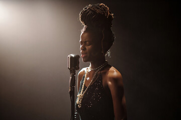 Black young woman singing into vintage microphone on stage with eyes closed wearing jewelry and...