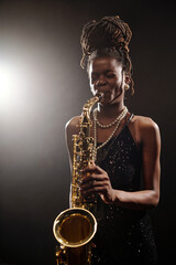 Black young female musician playing saxophone on stage, wearing jewelry and dress, eyes closed...