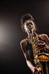 Black young adult woman playing saxophone on stage, wearing sleeveless top, standing under stage...