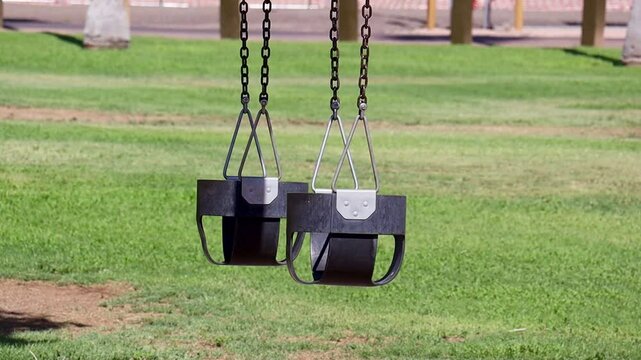 View of sunlit two black toddler swings hanging above well-kept green grass at Falcon Field Park