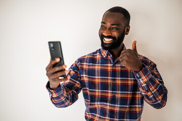 Ecstatic black man in cozy shirt standing in front of wall and having video call on smartphone.