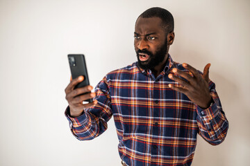 Angry black man in cozy shirt standing in front of wall and having video call on smartphone. He is arguing with someone.
