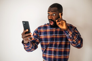 Surprised and shocked black man in cozy shirt standing in front of wall and having video call on smartphone.
