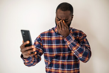 Tired black man in cozy shirt standing in front of wall and having video call on smartphone.