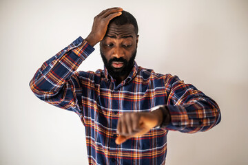 Anxious black man in cozy shirt standing in front of wall and looking at his wristwatch. He will be late.