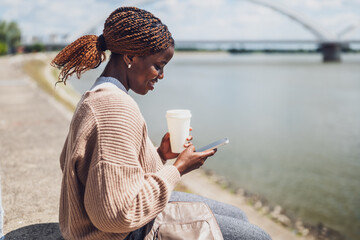 Young black woman is using smartphone and drinking coffee to go, outdoor by the river.