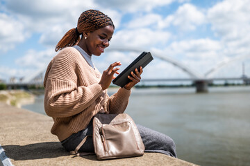 Young black woman is relaxing outdoor by the river. She is using digital tablet.