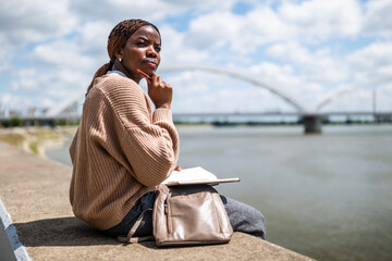 Portrait of thoughtful young black woman who is sitting by the river in the city and writing in note pad.