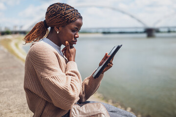 Young worried black woman is using digital tablet. She is sitting by the river in the city.