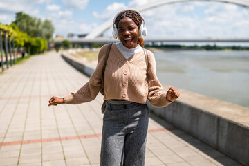 Happy young black woman is walking on foot path, listening music and smiling. She is enjoying sunny day outdoor.