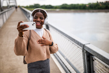 Happy young black woman is looking at her phone and smiling. She is enjoying sunny day outdoor and listening music while using smartphone.
