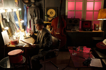 Middle aged Caucasian man wearing suit and hat sitting at dressing table holding glass and reading...