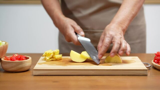 Close-up of Asian man's hands slicing potatoes on a wooden cutting board in the kitchen, preparing for cooking.