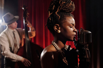 Black young adult woman singing into vintage microphone with eyes closed, wearing pearl necklace and gloves, Caucasian middle aged man playing double bass in background on stage