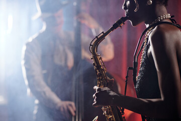 Black woman playing saxophone in foreground, wearing jewelry, with blurred Caucasian man playing...