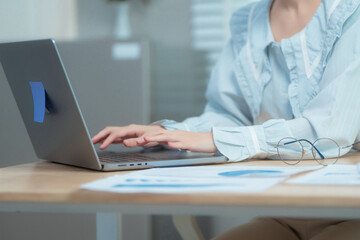 Confident beauty Asian office woman in a formal blouse hard working at her desk, reviewing a document with focus and a pen in hand. The workspace includes charts, a laptop, and a desktop computer.