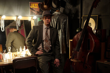 Caucasian middle aged man wearing fedora sitting beside double bass in jazz dressing room, vintage...