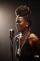 Black young woman singing into vintage microphone on stage, wearing elegant jewelry, eyes closed,...