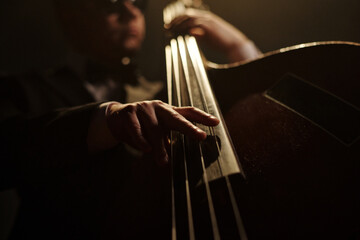 Middle aged Caucasian man playing double bass on stage, close up of hand plucking strings, jazz musician performing live music, blurred face in background, low angle view