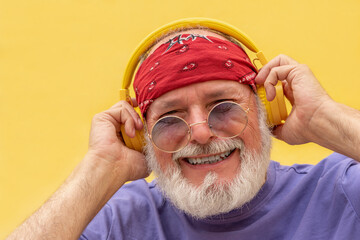 Happy senior man wearing bandana listening music with headphones and smiling on yellow background