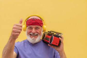Happy senior man listening to music with headphones and boombox giving thumb up on yellow background
