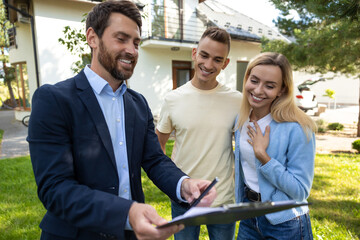 Real estate agent presenting clipboard with documents to couple in front of residential property