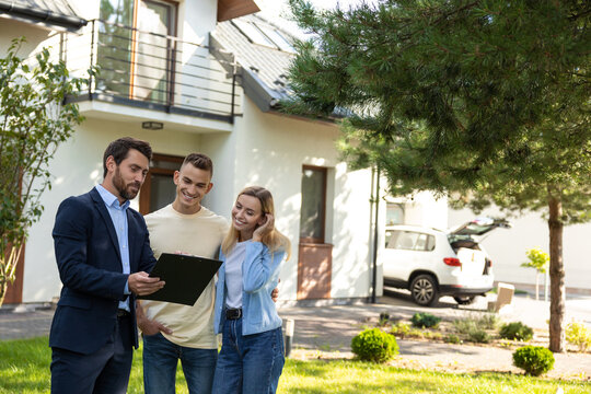 Real estate agent presenting clipboard with documents to couple in front of residential property
