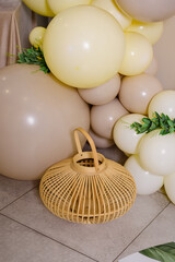 A decorative balloon arrangement in beige, yellow, and white tones, accented with green foliage, complementing a natural woven lantern on the floor.