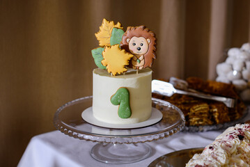 An adorable white first birthday cake featuring a green "1" and topped with a charming hedgehog cookie and autumn leaves, displayed on a glass cake stand