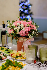 A beautifully decorated table featuring a vibrant pink floral bouquet, appetizers, candles, and holiday ornaments, with a blurred Christmas tree in the background.