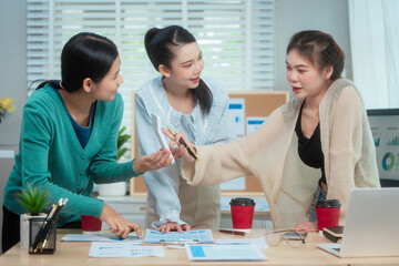 Three Asian women collaborate at shared office desk filled with documents, charts, coffee. One...