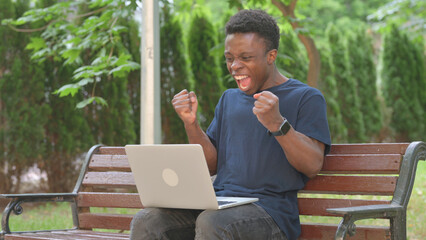 Cheerful Casual African Man with Laptop Enjoying Win on Park Bench