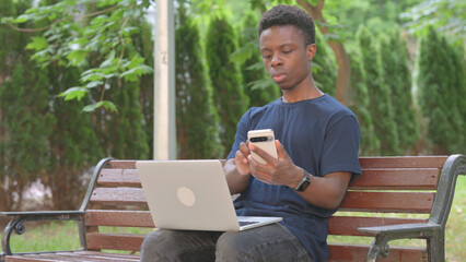 Casual African Man Using Smartphone and Laptop Outdoors