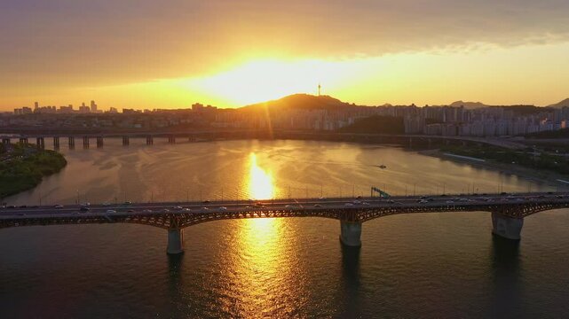 Scenic sunset view of Seoul city with cars crossing the bridge over Han River, urban skyline and Namsan Seoul Tower in the background.
