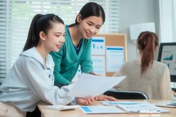 Three Asian women collaborate at shared office desk filled with documents, charts, coffee. One...